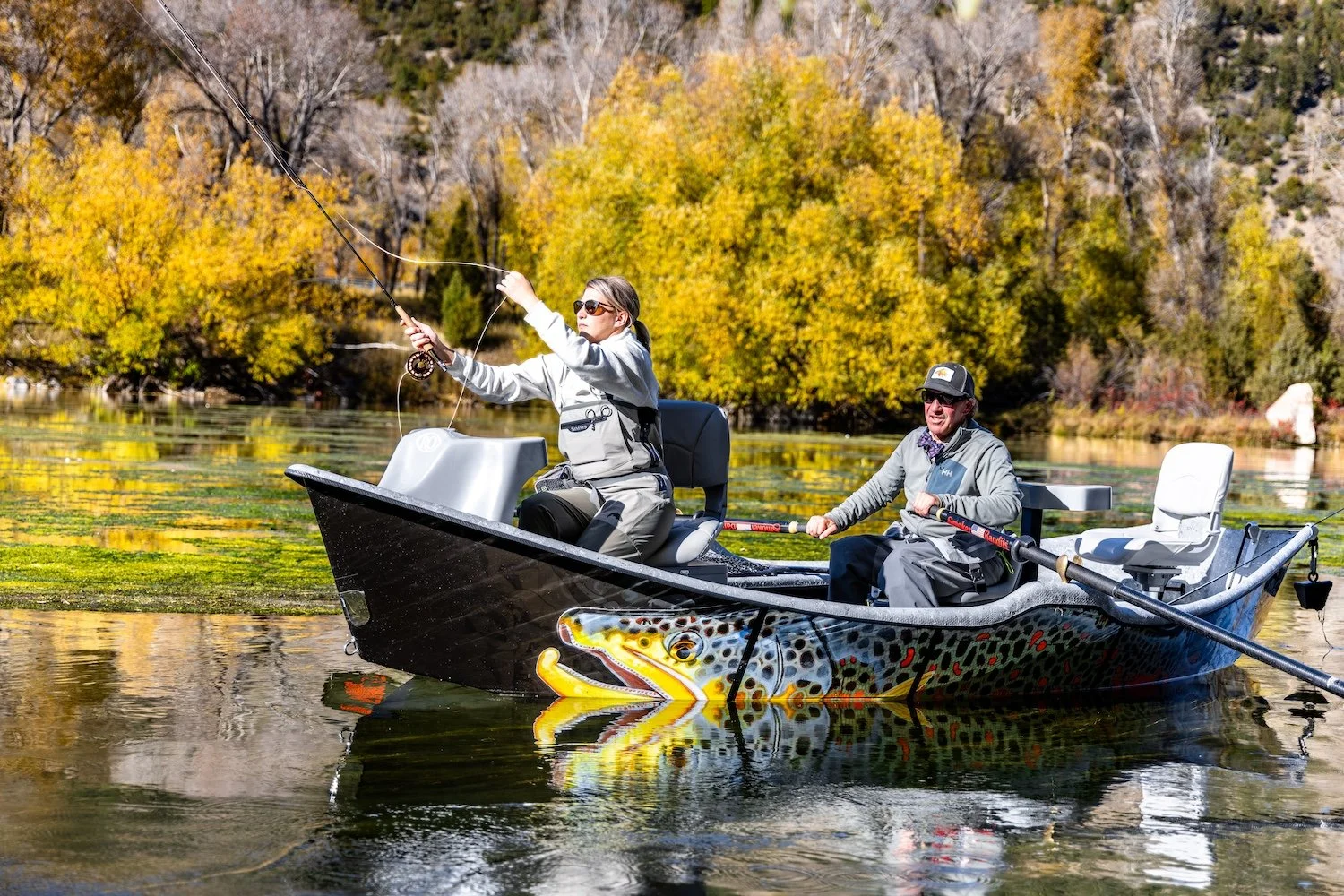 Two people fishing from a small boat on a lake with autumn-colored trees in the background. Livingston, Montana, USA