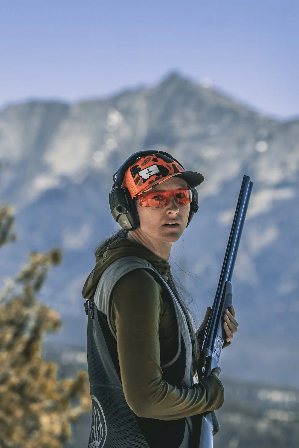 A woman wearing a black vest, orange camouflage cap, orange tinted glasses, and hearing protection, holding a shotgun, with snow-covered mountains in the background. Big Sky, Montana, USA