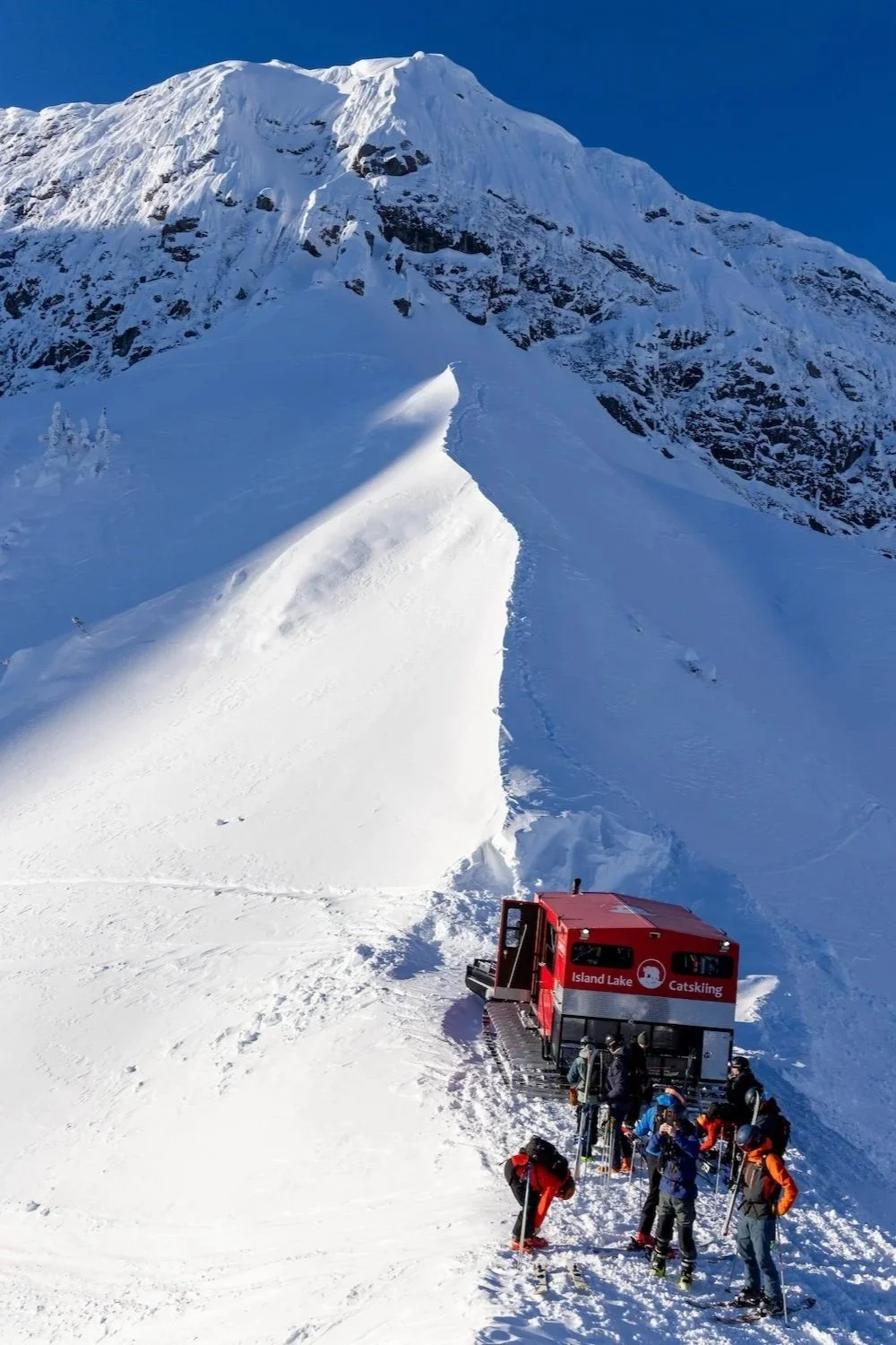 Group of people preparing for skiing near a snowcat vehicle on a snowy mountain slope, with a large snow-covered mountain in the background.