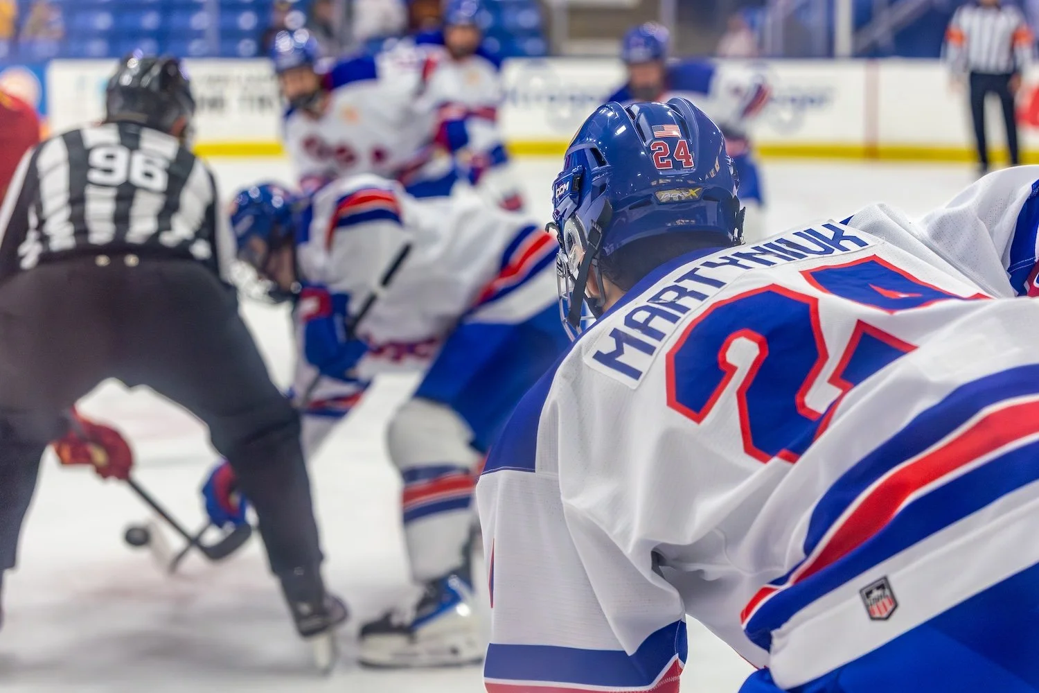Hockey players face off during a game on ice, with a referee in black and white nearby.