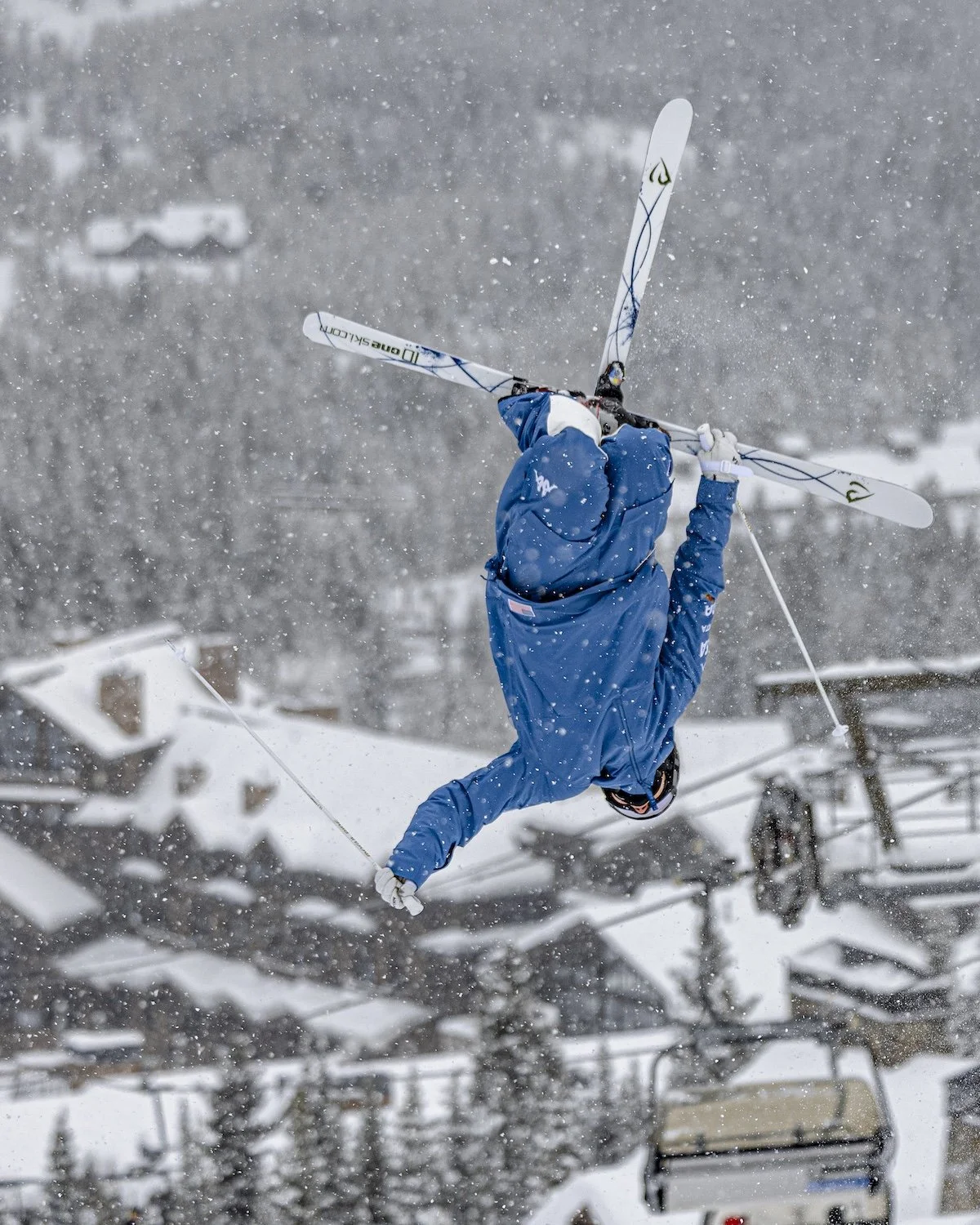 A skier in mid-air performing a flip on a snowy mountain, wearing a blue jacket, black helmet, and white gloves, with ski lift chairs and snow-covered trees in the background during snowfall.