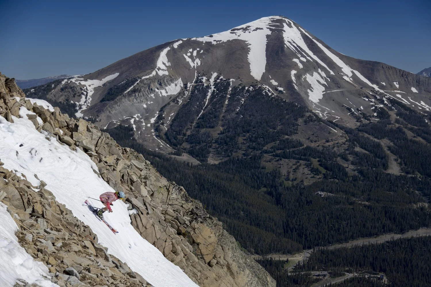 A skier in pink gear climbing a steep snow-covered slope on a rocky mountain with a forested mountain range and a snow-capped volcano in the background.