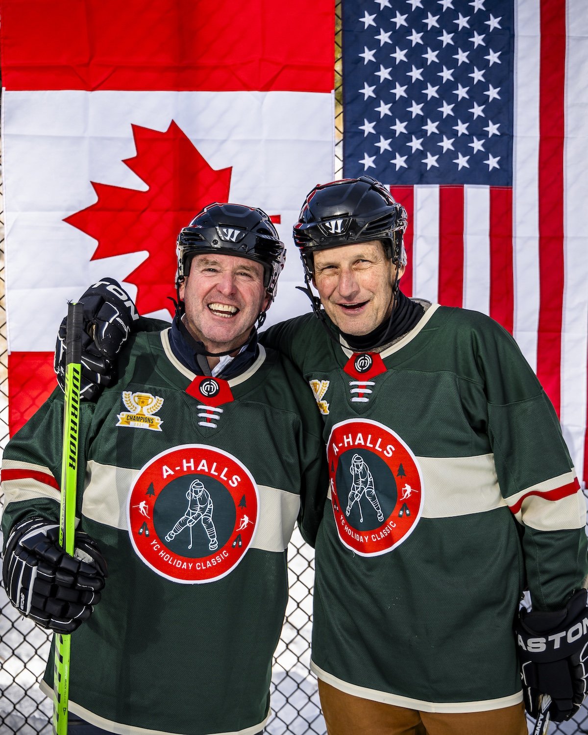 Two men in hockey jerseys smiling and posing with arms around each other, standing in front of Canadian and American flags.