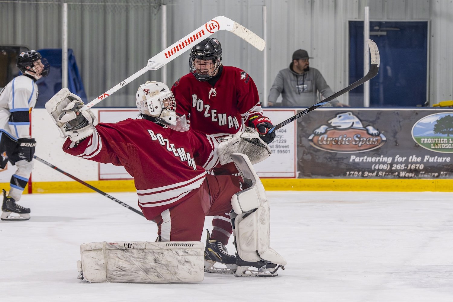 Ice hockey goalies in red and white jerseys with 'Bozeman' written on them, one in a crouched position and the other standing, on the ice rink. A player in white skates nearby, and a man watches from behind the rink boards.