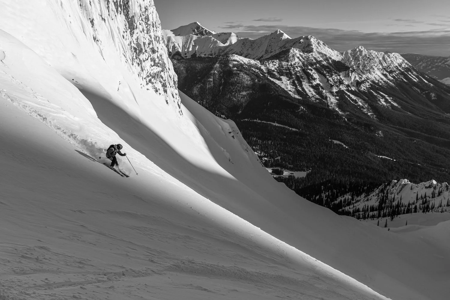 A skier descending a steep snow-covered mountain slope with a mountainous landscape in the background, black and white photo.