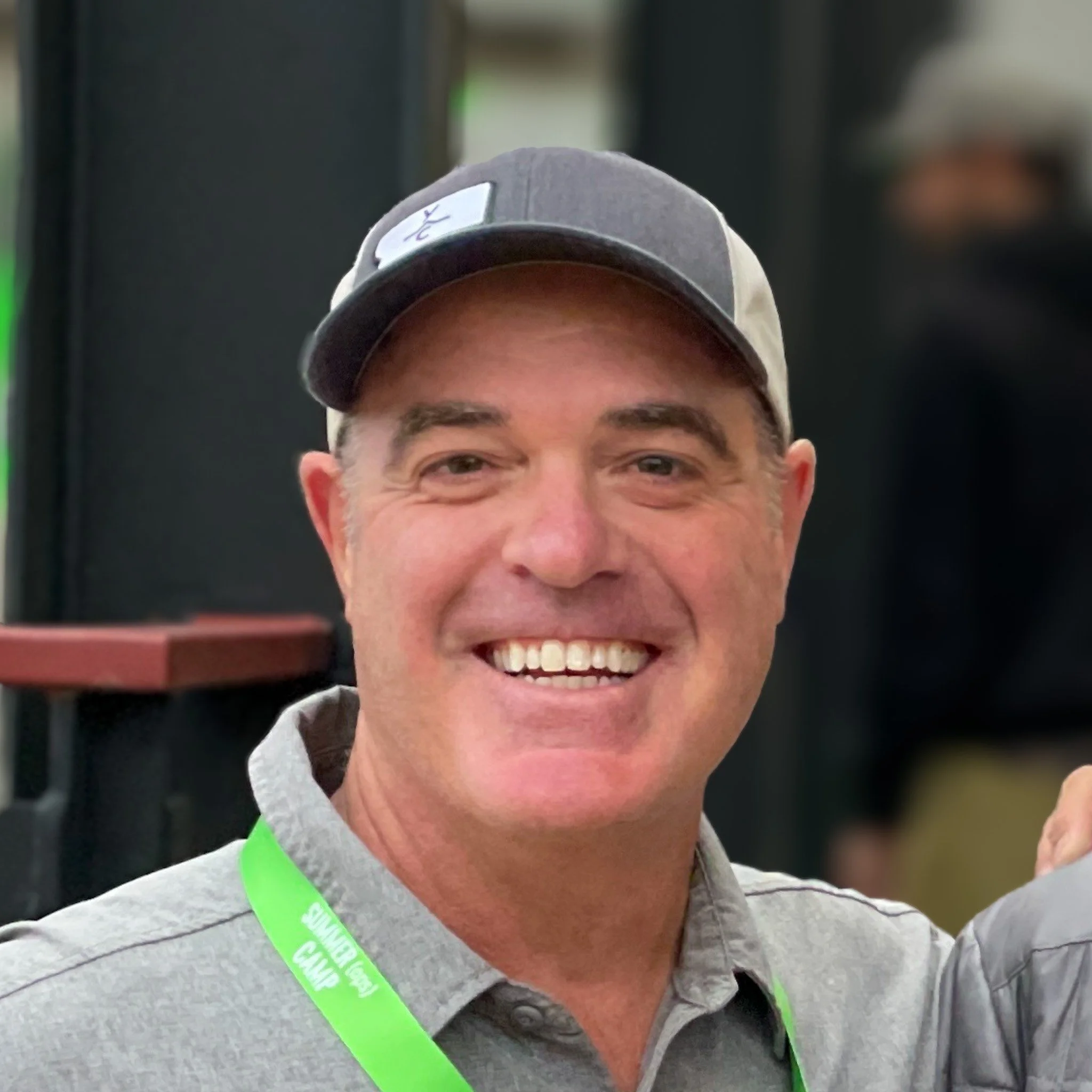 A young man wearing a grey hoodie and a grey baseball cap with a tag, smiling and holding a smartphone in front of him taking a selfie, with a blurred background.