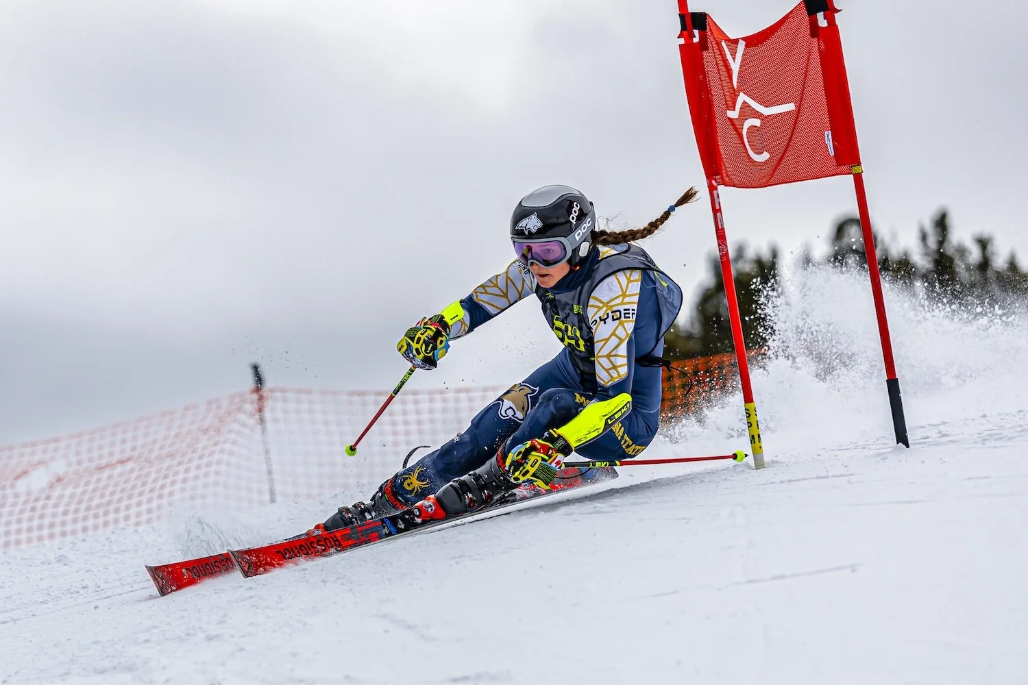A female alpine skier in a blue and yellow racing suit navigating a slalom race on a snowy slope, wearing a helmet and goggles, with a red gate and safety net in the background.