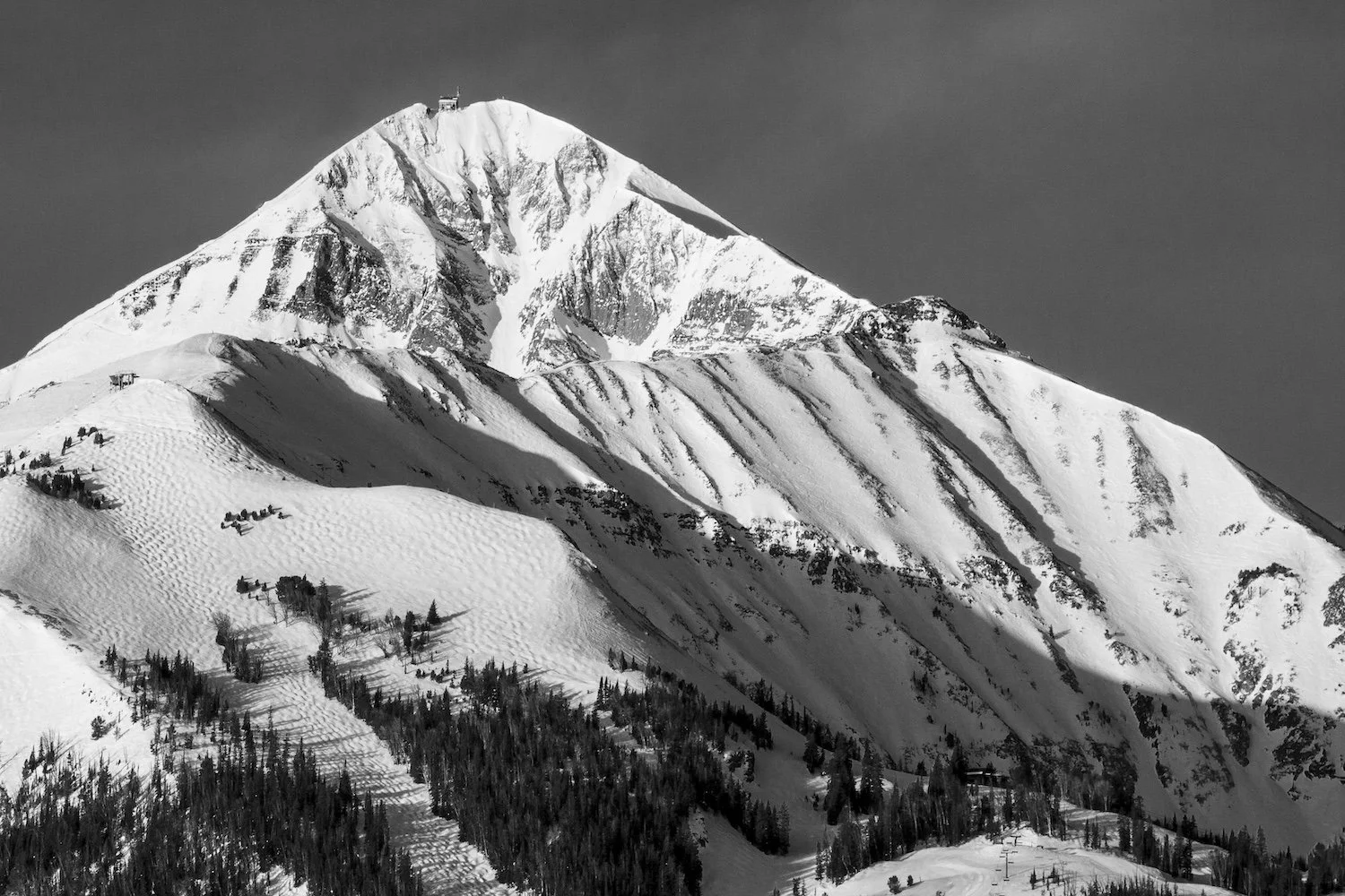 Black and white photo of a snow-covered mountain peak with a structure at the summit and ski slopes on the hillside. Lone Peak, Big Sky, Montana, USA