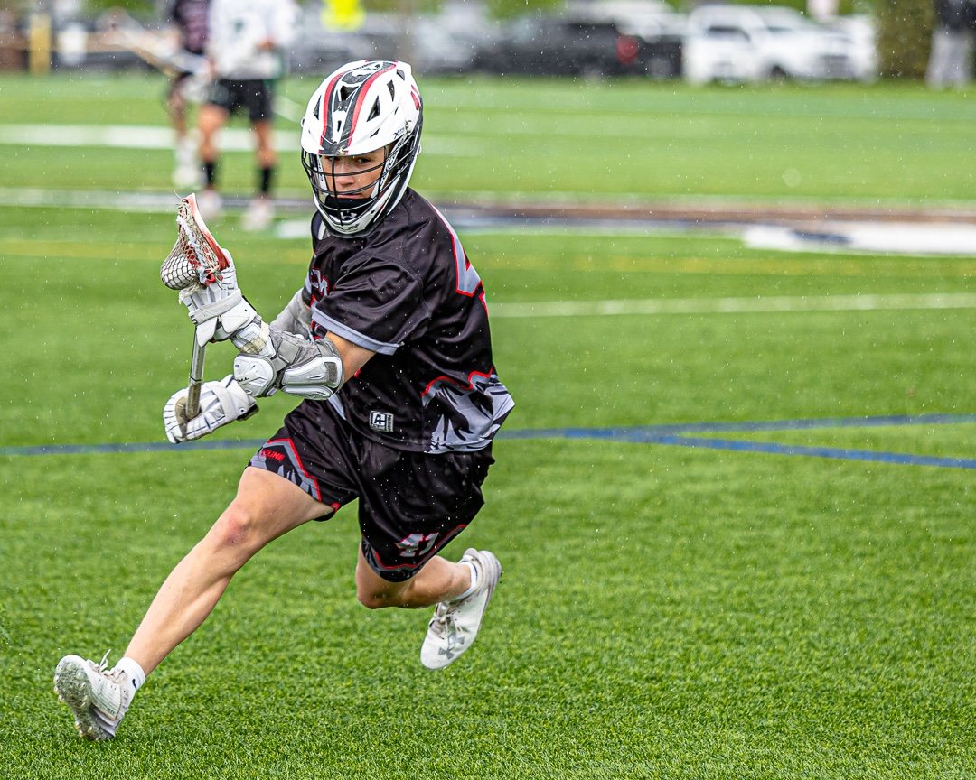 A young lacrosse player in black gear running on a wet green field during a rainstorm, wearing a helmet and holding a lacrosse stick.