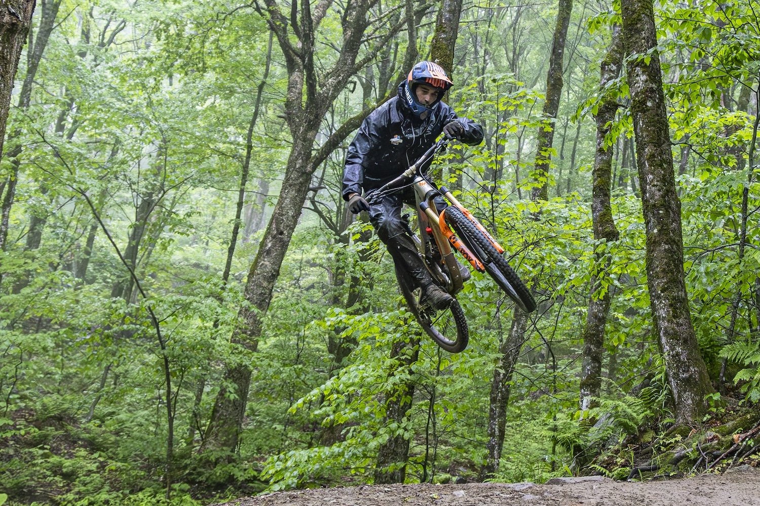 A mountain biker riding through a forest, airborne over a dirt trail on a rainy day. Killington, VT, USA