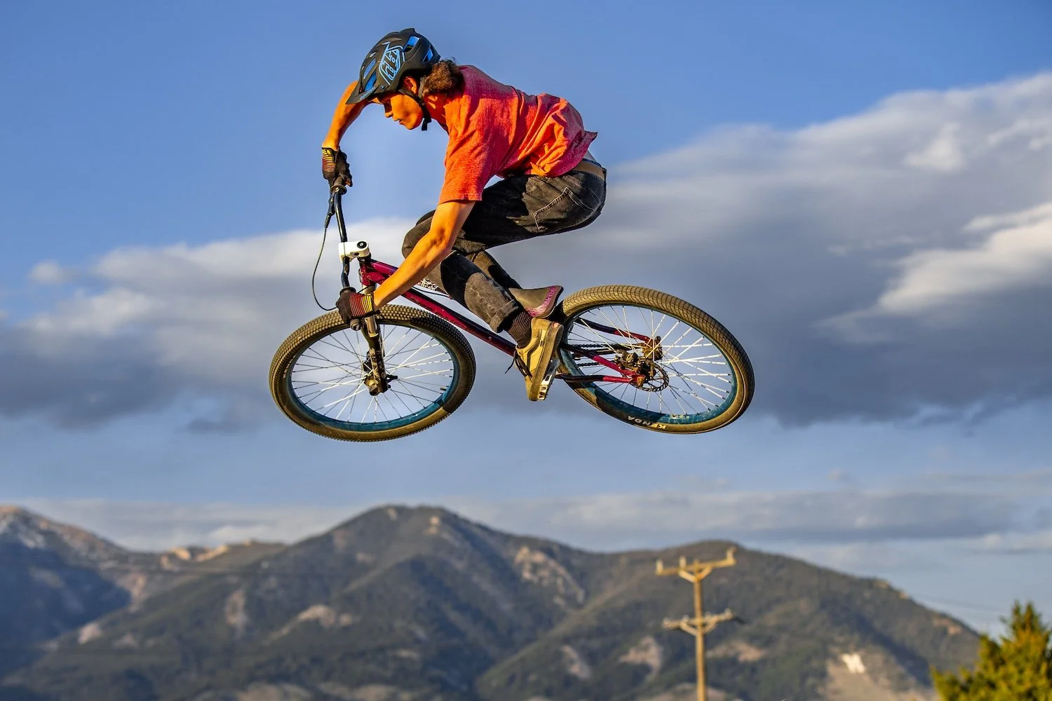 Person in mid-air performing a bike jump against a background of mountains and blue sky. Bozeman, MT, USA