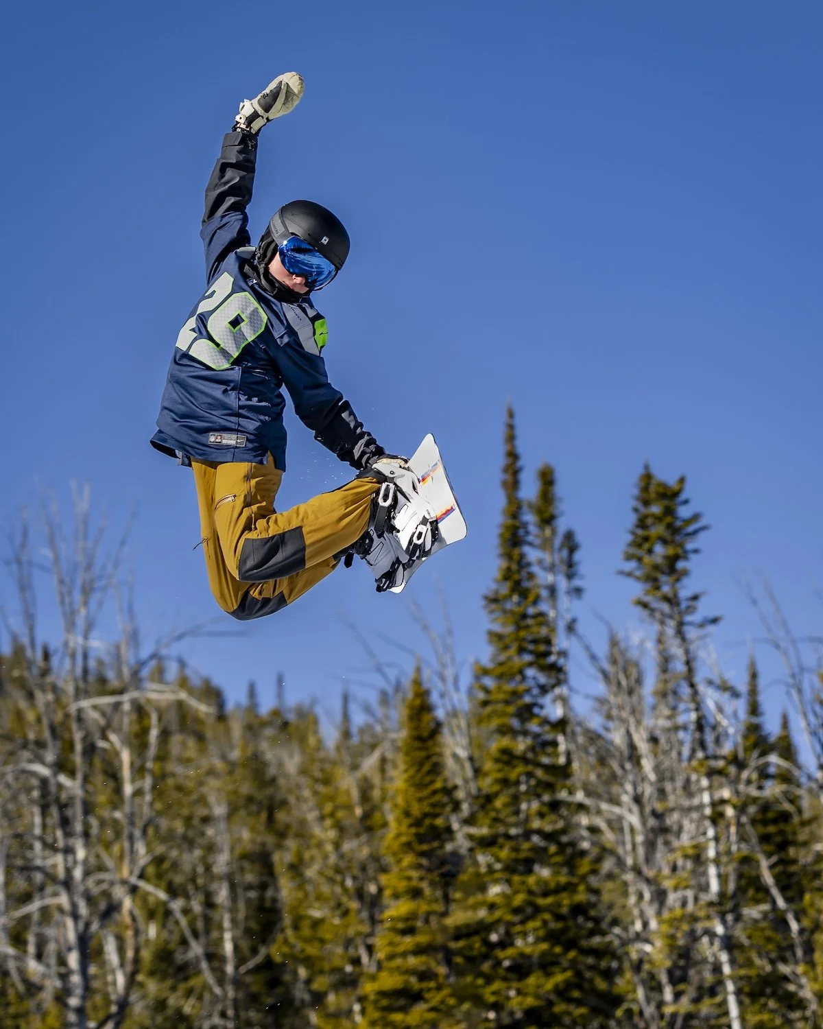 A person wearing a helmet, goggles, and winter gear is mid-air jumping on a snowboard against a clear blue sky with trees in the background.
