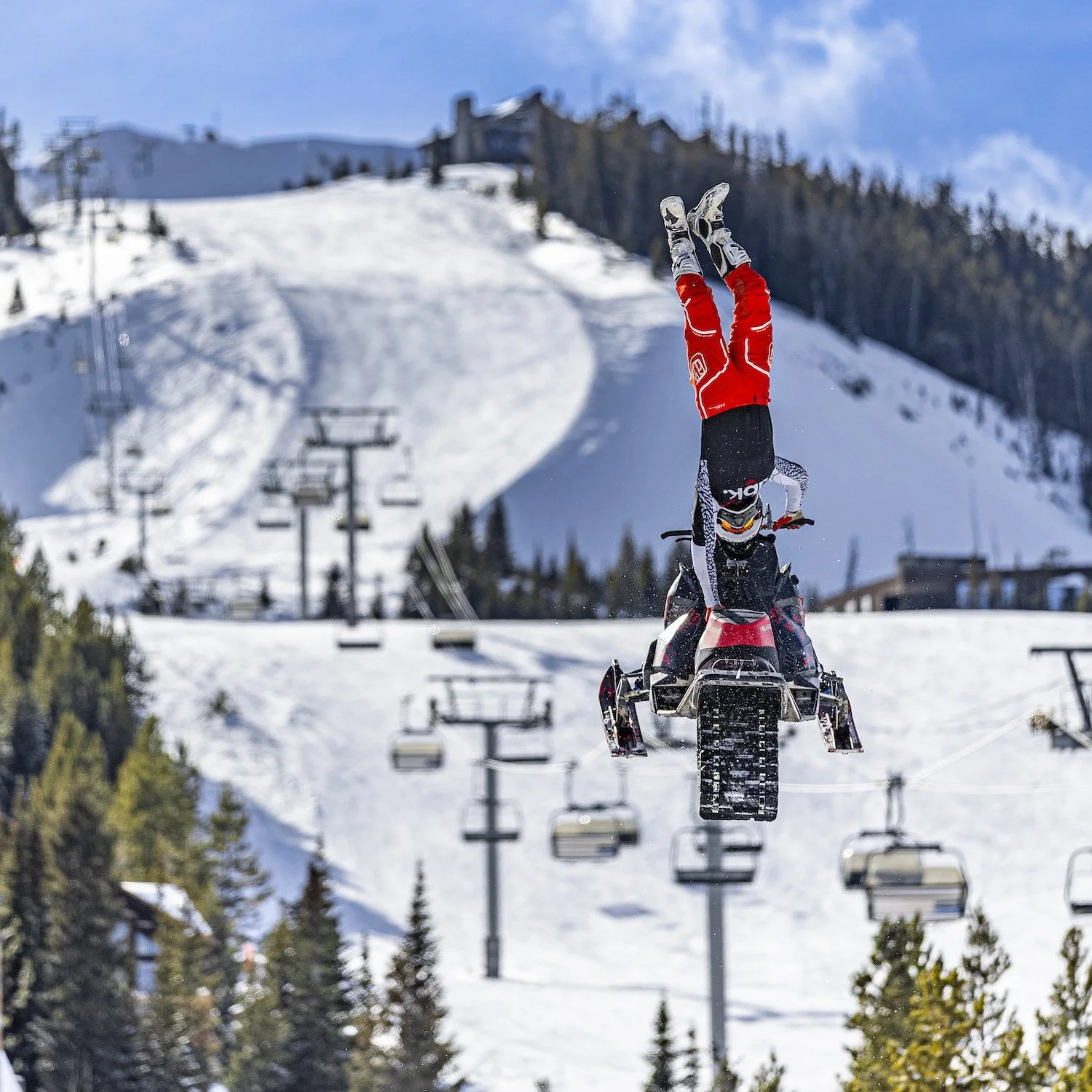 A person doing a headstand on a snowmobile at a ski resort, with ski lifts and snow-covered slopes in the background. Freestyle snowmobile. Freestyle snowmobile show, Kieth Sayers 
