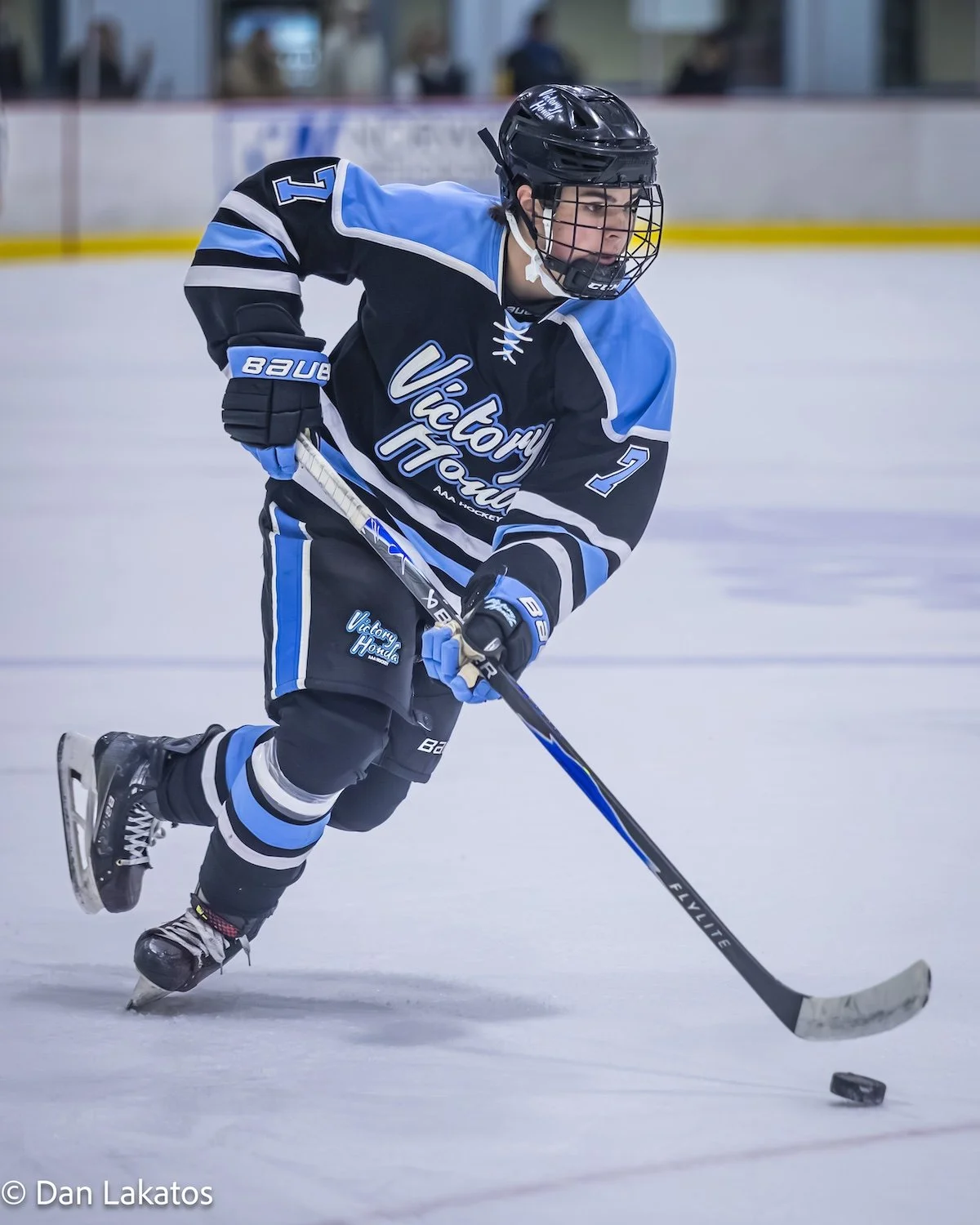 An ice hockey player in a blue, black, and white uniform with the words "Victory Honda" on it, skating on the ice and handling a hockey puck with his stick, wearing a black helmet and gloves.