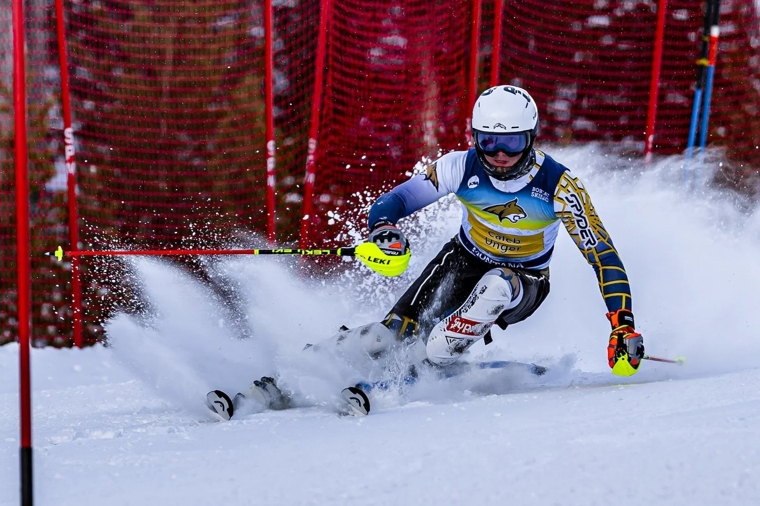 An alpine skier in a blue, white, and black uniform skiing on snow towards a red gate on a ski course with red safety netting in the background.