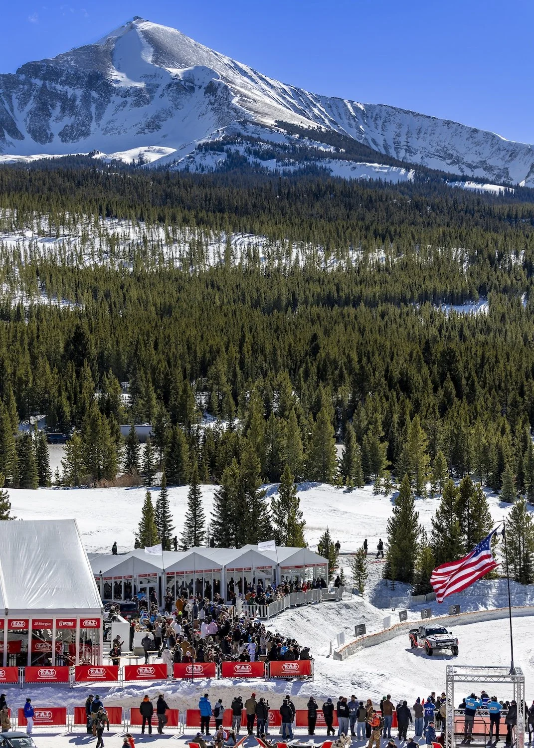 Crowd gathered at a car race with a large snow-capped mountain in the background, a dense pine forest, and tents set up along the snow. An American flag is flying near the event. F.A.T. ICE RACE 2026, Big Sky, Montana USA. Dan Lakatos Photo