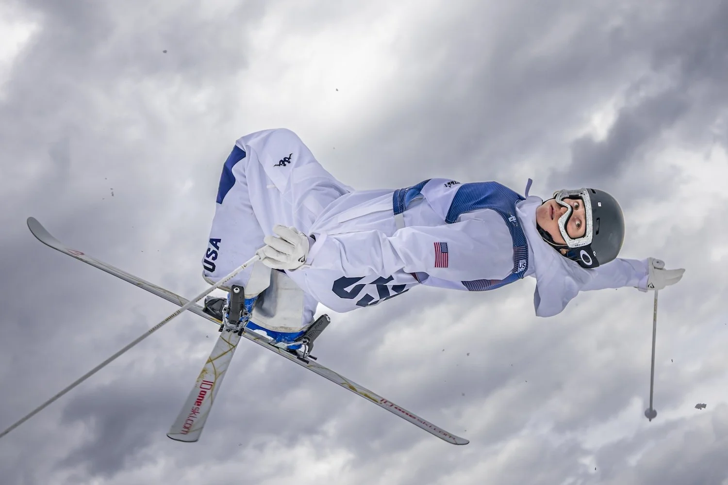 A skier wearing a white USA team uniform and black helmet, mid-air in a ski jump against a cloudy sky, holding ski poles.