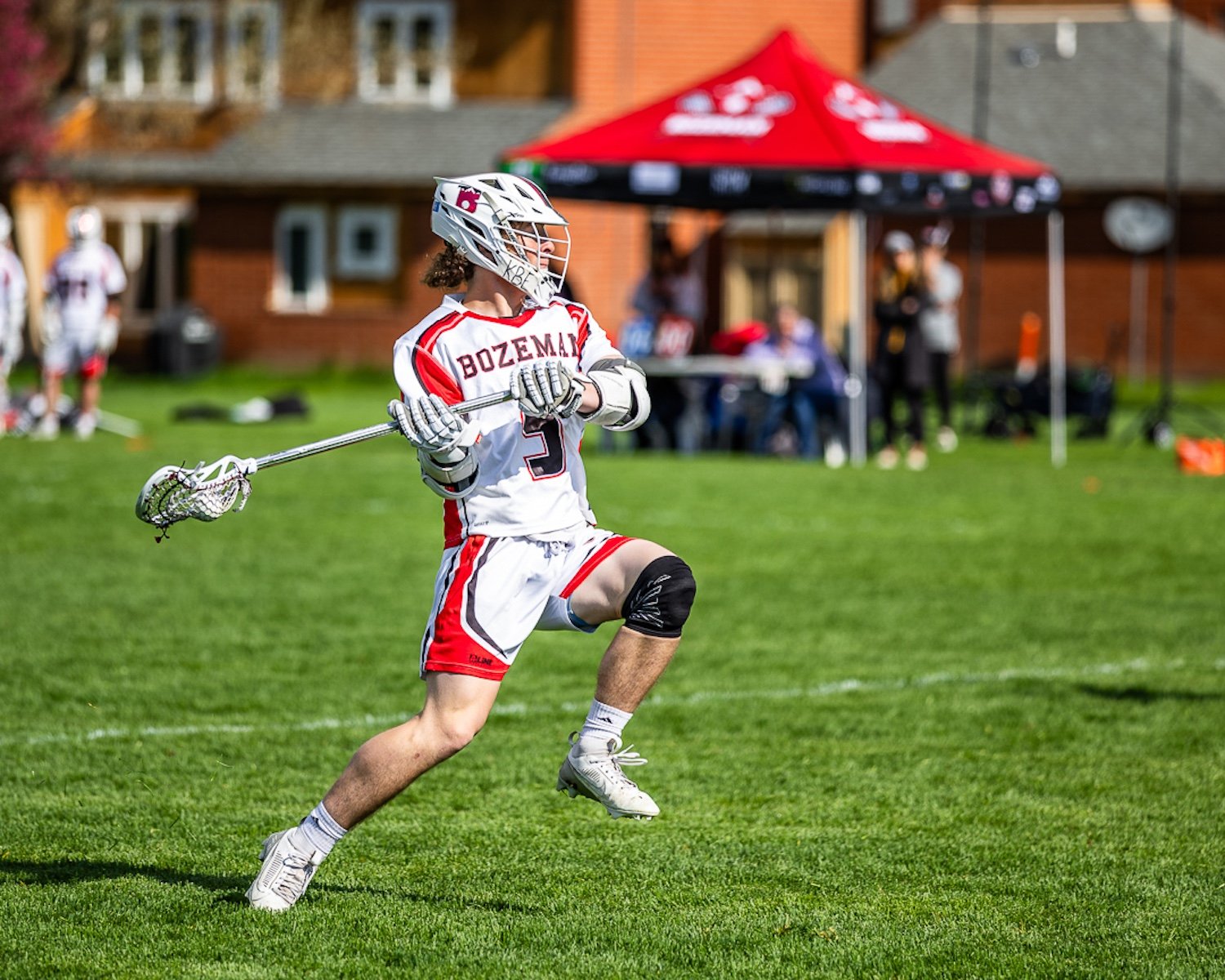 A young male lacrosse player in white and red uniform with 'Bozeman' on the jersey, running on a grassy field holding a lacrosse stick, with a red tent and other players and spectators in the background.