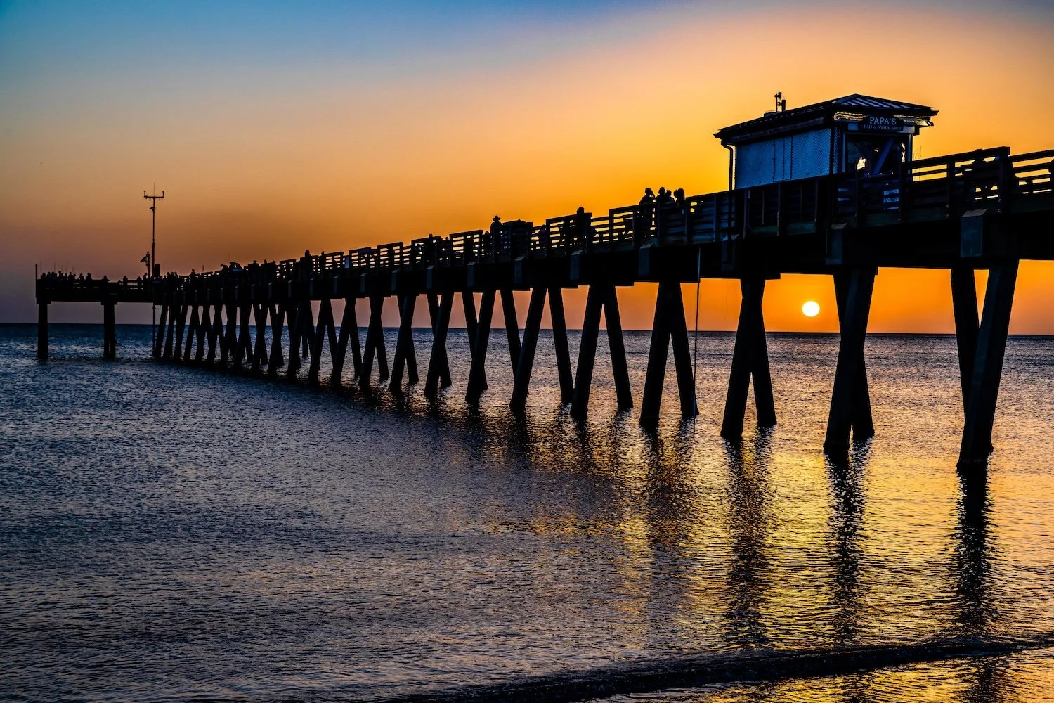 Silhouette of a pier extending over the water at sunset, with people and a small structure on the pier, and the sun near the horizon in the background. Venice Beach, Florida
