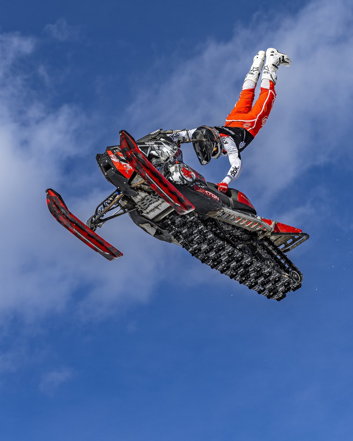 A person performing a snowmobile stunt mid-air against a blue sky, wearing a black and white helmet and red snowmobile gear. Freestyle snowmobile show, Kieth Sayers 