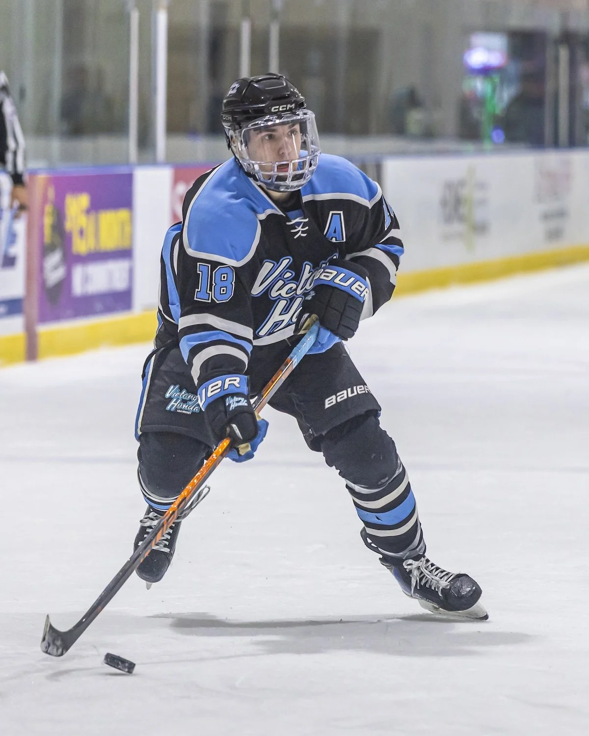 An ice hockey player in black and blue jersey with the number 18, wearing a black helmet and gloves, skating on the ice rink with a hockey stick and puck.