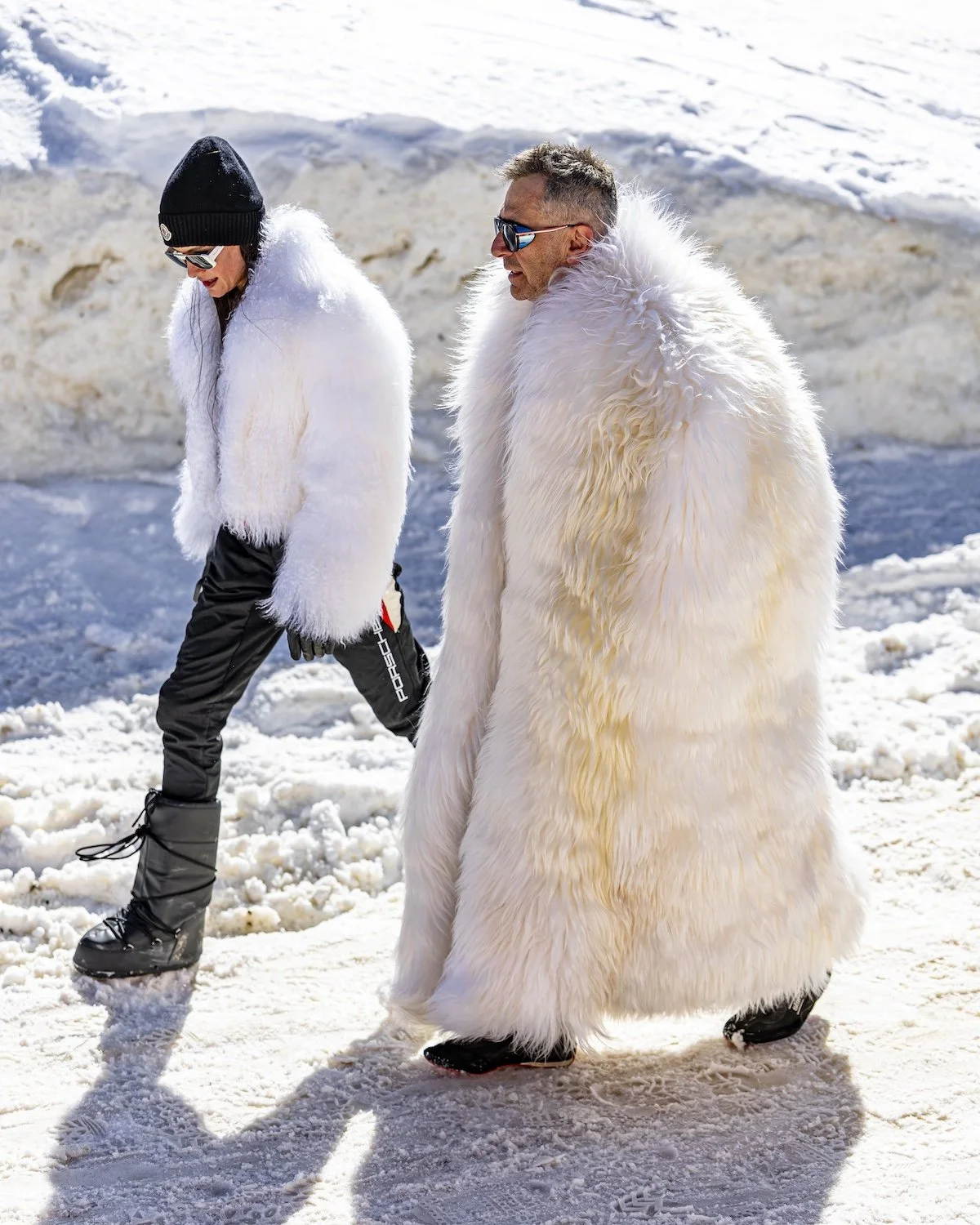 Two people walking in snowy environment, both wearing large white fur coats, black snow boots, sunglasses, and warm clothing, with snow and ice background. F.A.T. ICE RACE 2026, Big Sky, Montana USA. Dan Lakatos Photo