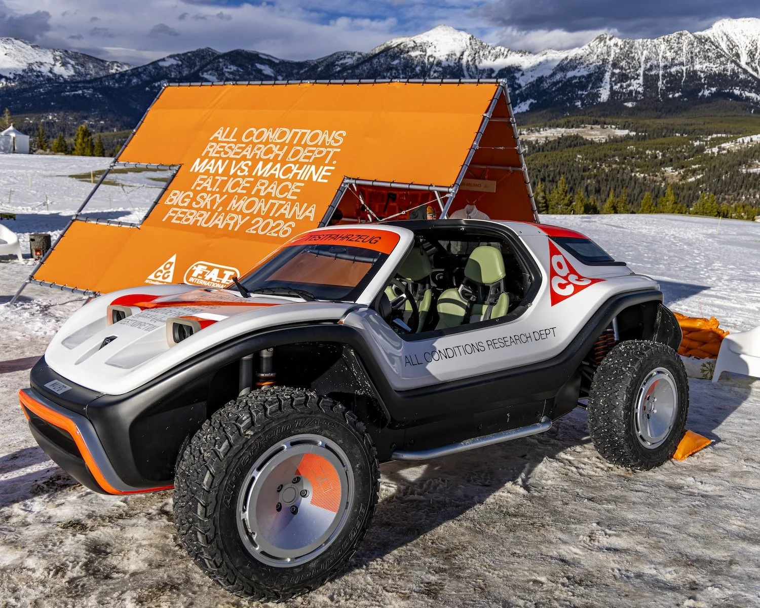 Off-road vehicle with a sleek design, black, white, and orange accents, parked on snowy ground with mountains in the background. F.A.T. ICE RACE 2026, Big Sky, Montana USA. Dan Lakatos Photo