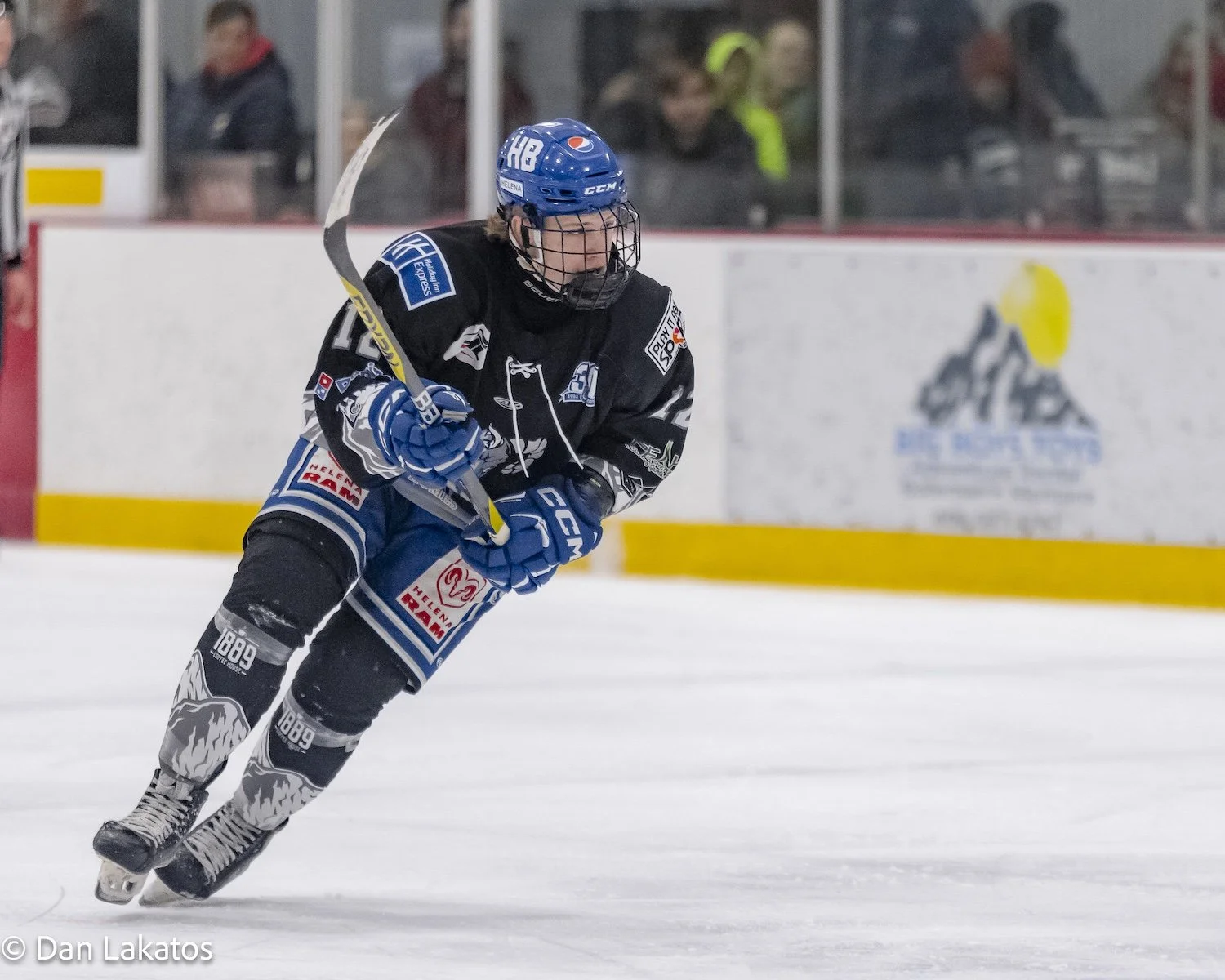 An ice hockey player wearing a blue helmet, black jersey with blue and white accents, and gloves skating on ice during a game.
