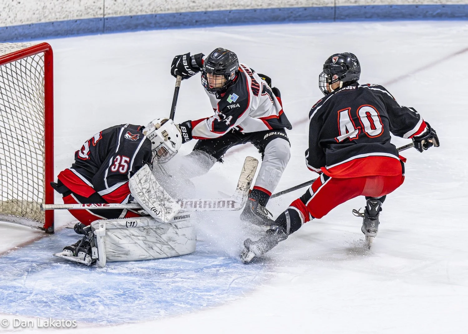 Ice hockey players in action near the goal, with the goalie in a crouched position blocking the puck, while two other players attempt to score or defend.