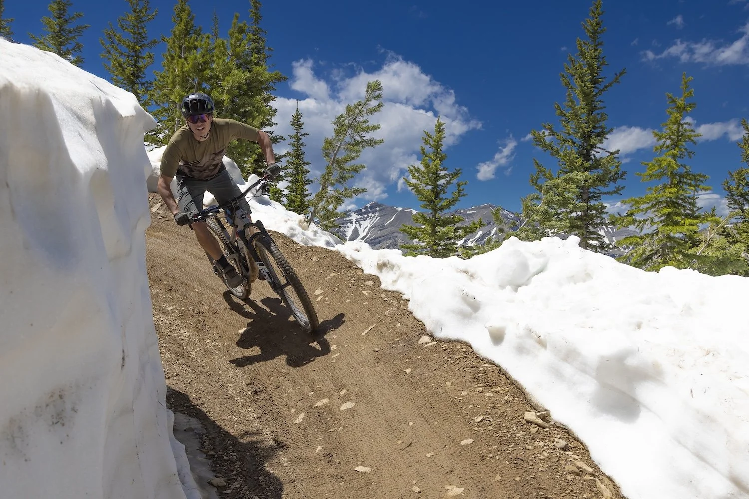 A person mountain biking on a dirt trail surrounded by snow and evergreen trees in a mountainous area under a blue sky with clouds. Big Sky, MT, USA