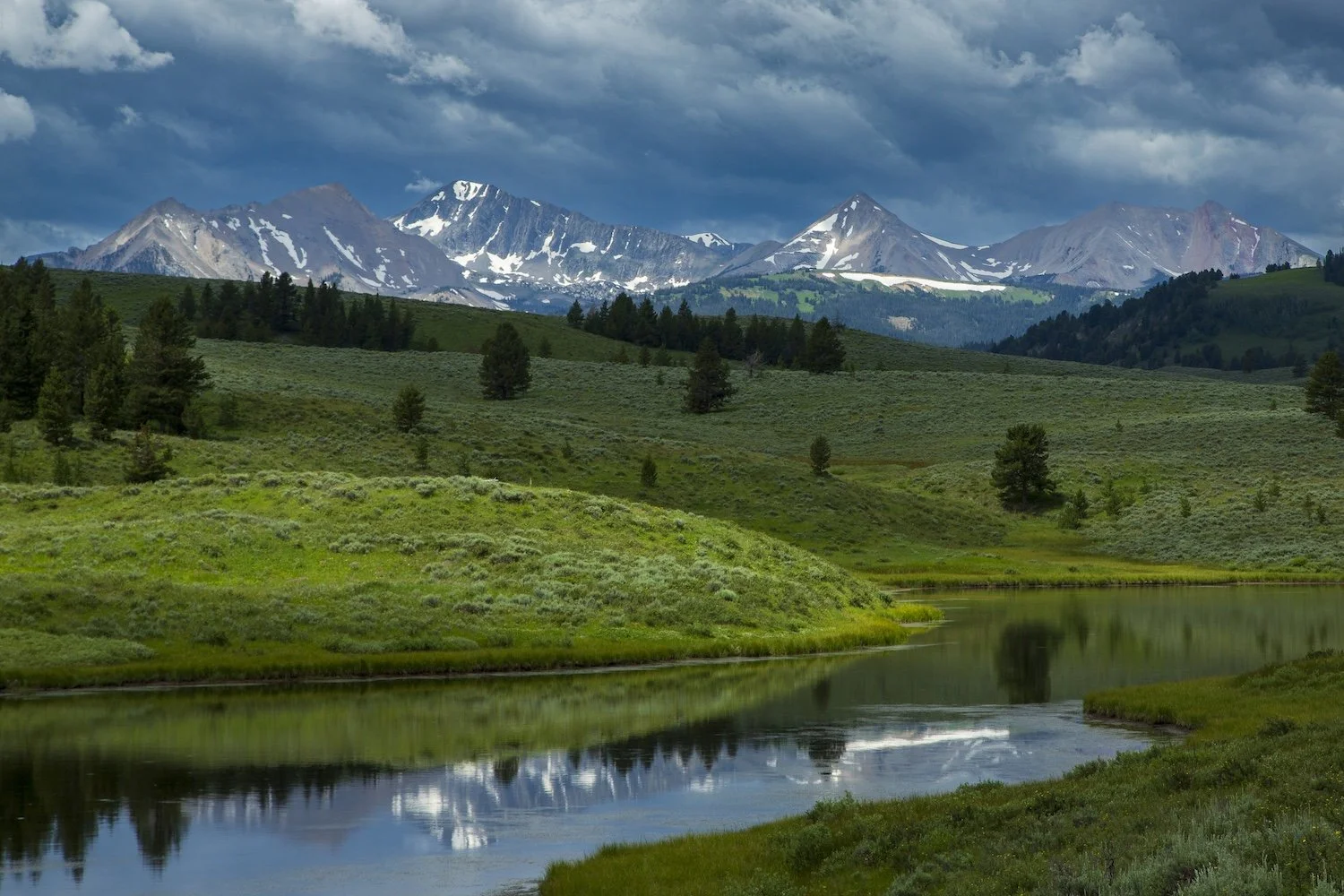 A scenic landscape featuring snow-capped mountains in the background, green rolling hills, trees, and a reflective river in the foreground under a cloudy sky. Madison Range near Big Sky, Montana, USA