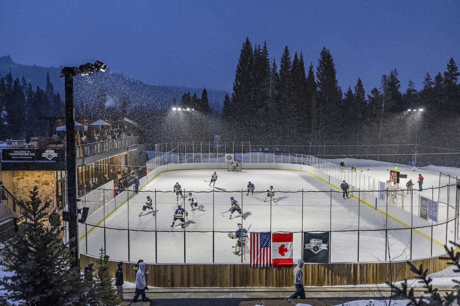 An outdoor ice hockey game taking place on an ice rink at dusk, surrounded by trees. Players are actively playing, with some skating and a goalie near the net. Spectators watch from the upper level, and banners with American and Canadian flags are di