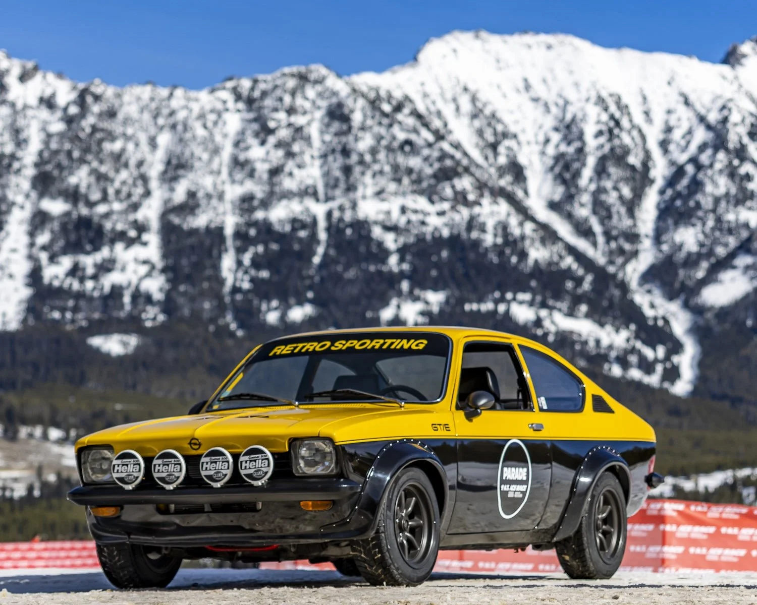 A vintage yellow and black racing car with four large front lights, parked on a gravel area with snow-capped mountains in the background. F.A.T. ICE RACE 2026, Big Sky, Montana USA. Dan Lakatos Photo