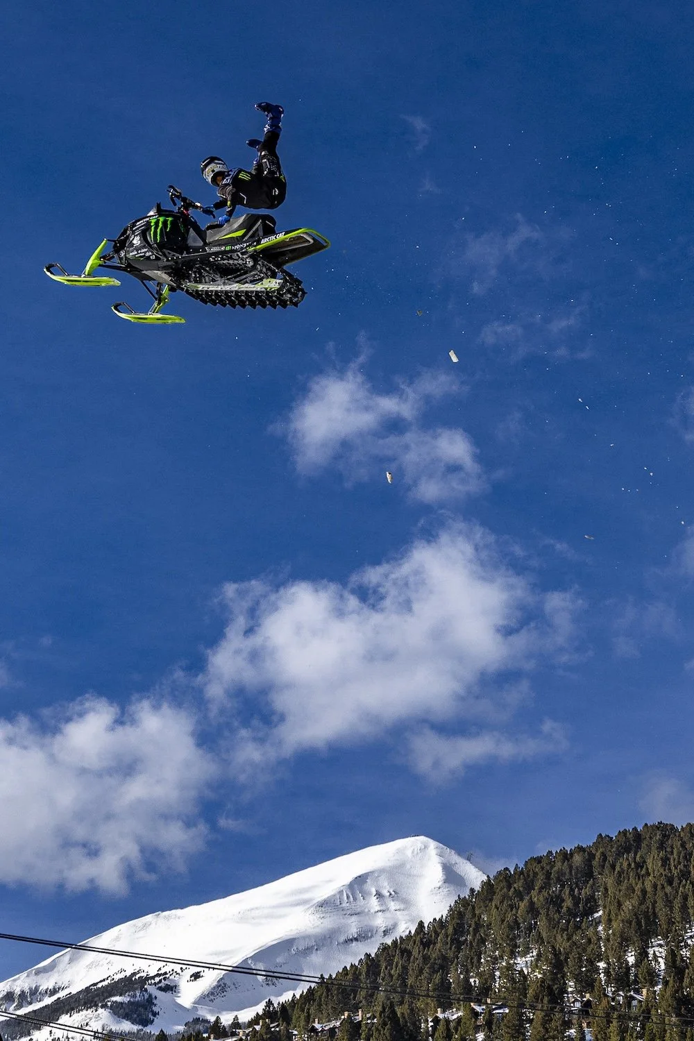 A snowmobile with a rider performing a stunt in the air above a snowy mountain and forest under a blue sky with clouds. Freestyle snowmobile show, Kieth Sayers 