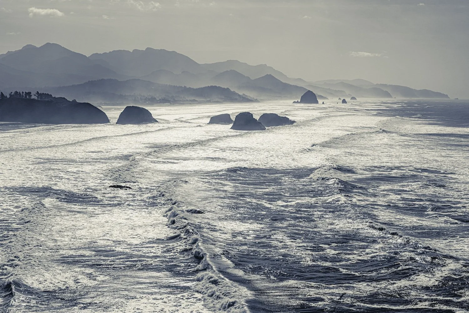 View of a rocky coastline and distant mountains with ocean waves and foam in the foreground.