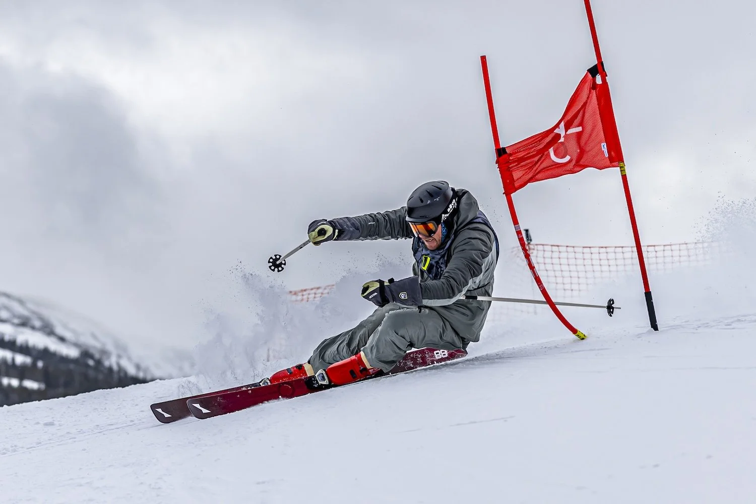 Skier in gray gear navigating through a slalom course on snow, with a deflected red gate in the background and snow-dusted mountains in the distance.