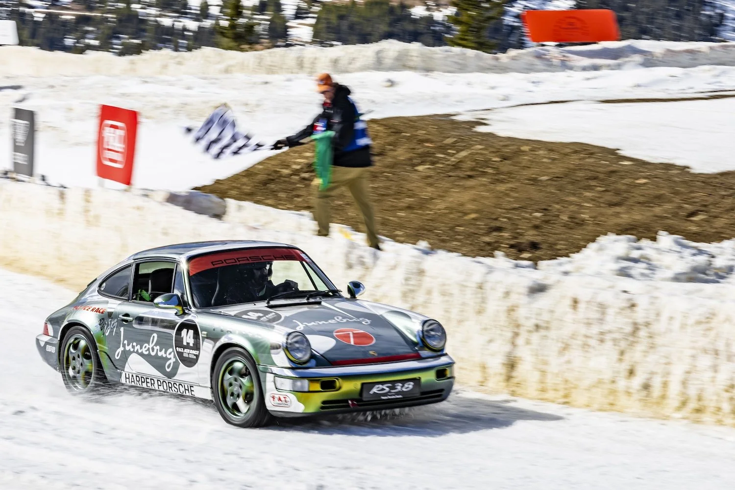 A vintage Porsche race car driving on a snow-covered track during a racing event, with a person standing on the side holding flags in the background.
