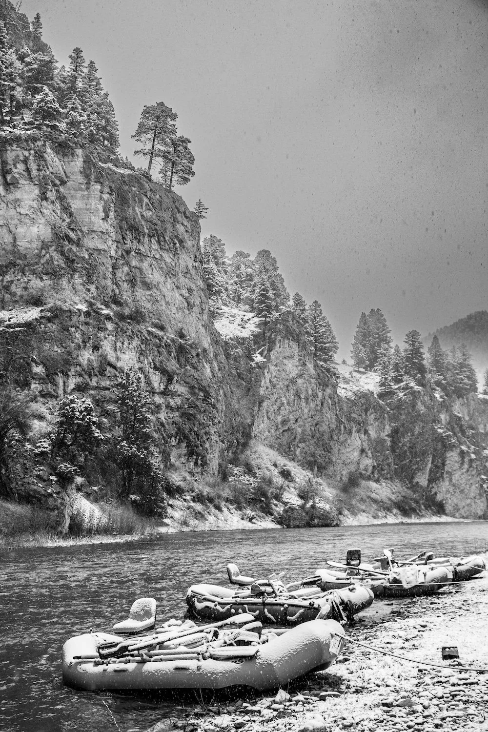 Snow-covered inflatable boats docked by a river with a snowy forested cliff in the background. Smith River, Montana, USA