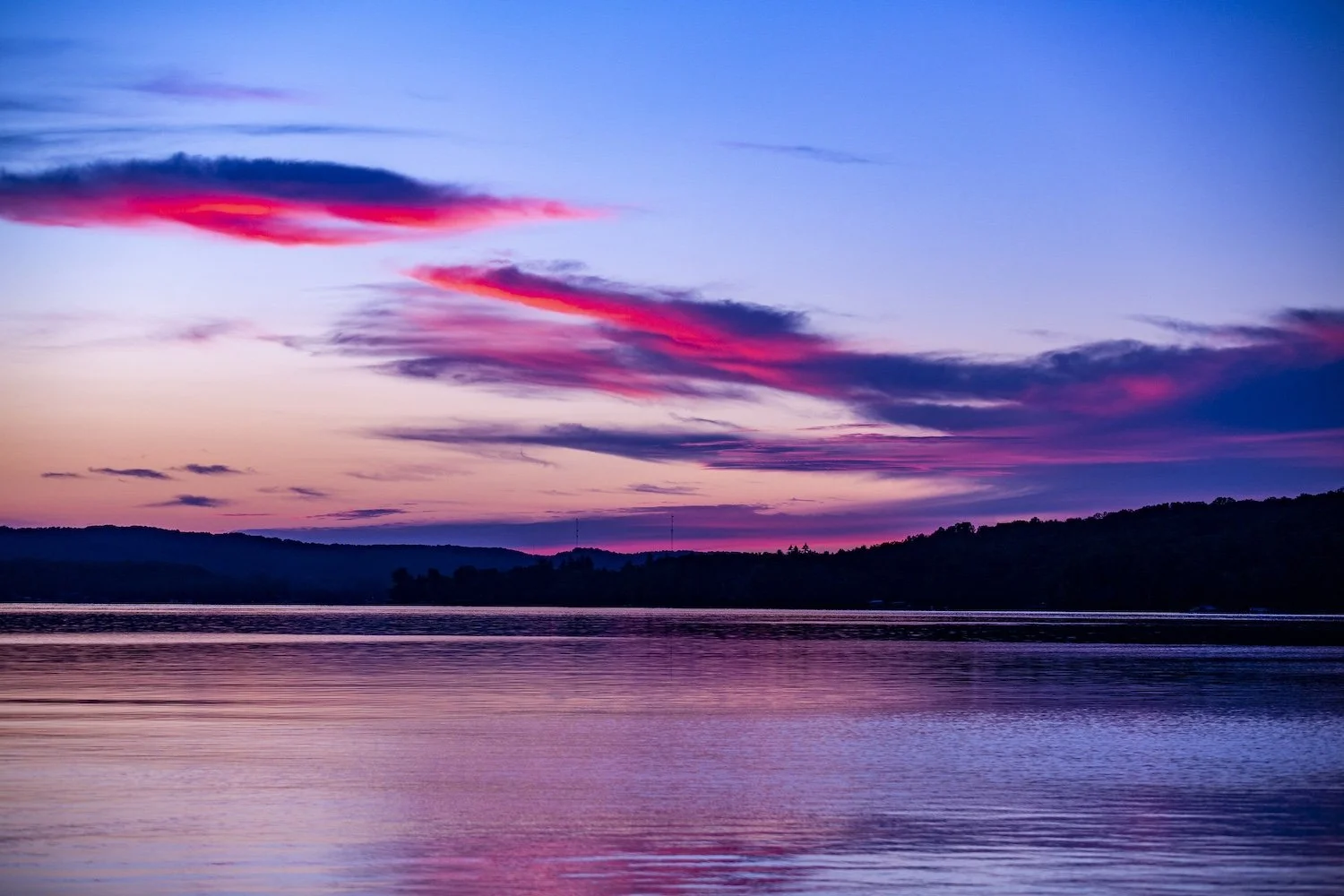 A colorful sunset over a calm body of water with reflections, silhouetted hills, and vibrant pink and purple clouds in the sky. Intermediate Lake, Bellaire, Michigan, USA