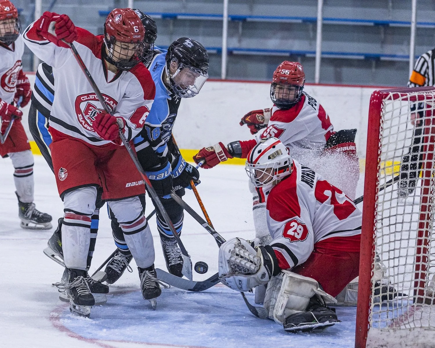Hockey players battling near the goal, with one goalie in white and red gear on his knees trying to block the puck, while opposing players in black and blue jerseys attempt to score.