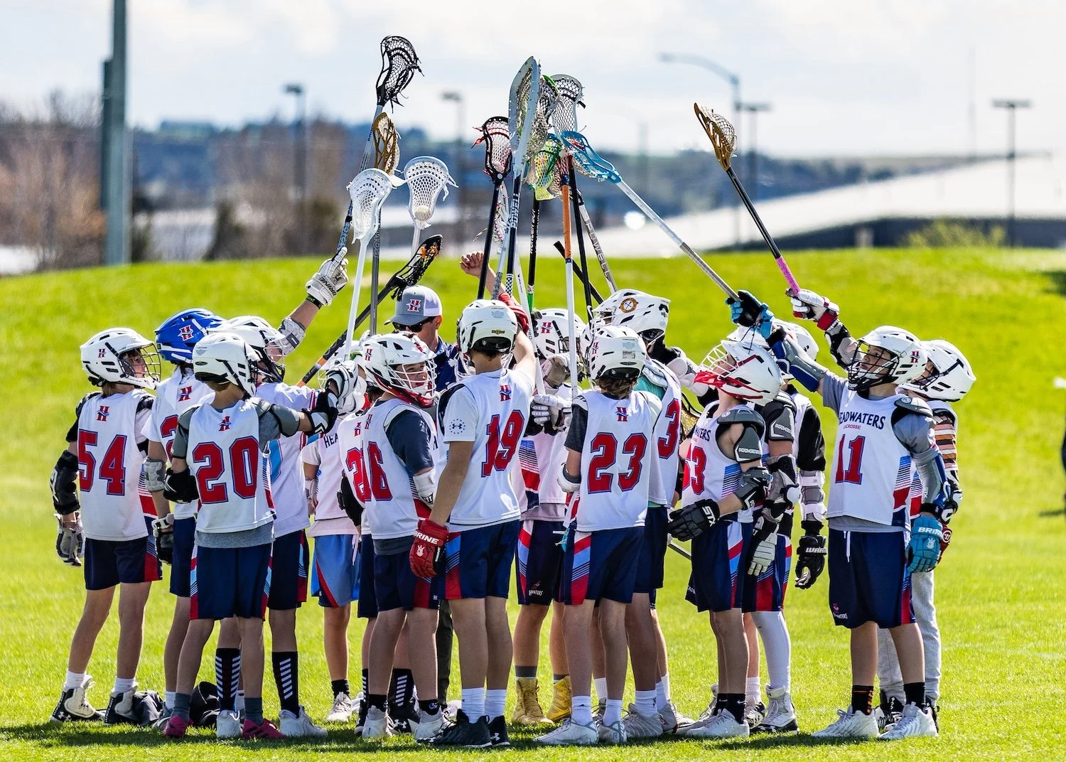 Youth lacrosse team wearing white jerseys and helmets huddled together on a field, raising their lacrosse sticks in the air during a game or practice.