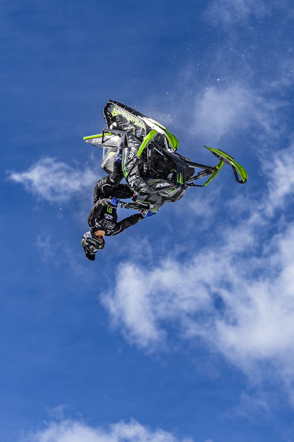Freestyle snowmobile rider flips mid-air on a green and black dirt bike against a blue sky with clouds. Freestyle snowmobile show, Kieth Sayers 