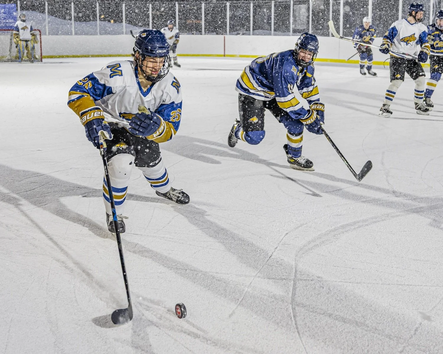 Hockey players in blue and white jerseys skating on ice during a game in an indoor rink, with some players in the background and snow falling outside.