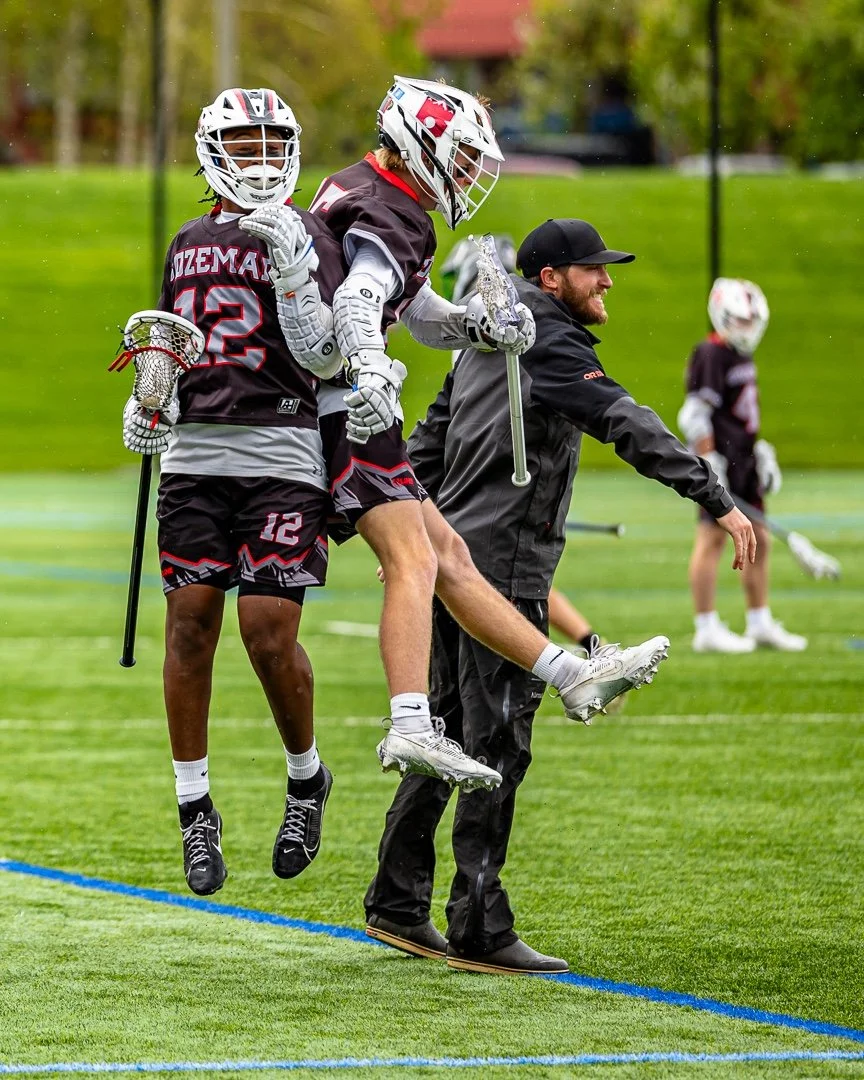 Lacrosse players in gear clebrating on a field with a coach.