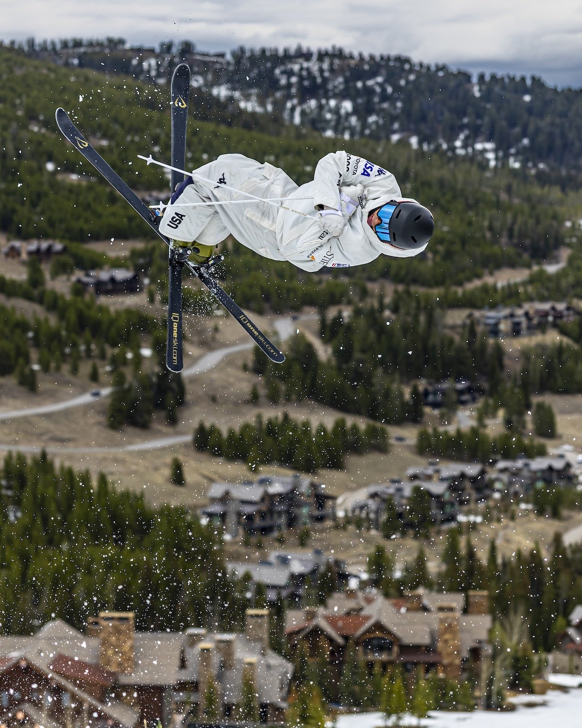A skier in white gear performing a jump in mid-air, with a mountainous landscape and houses below.