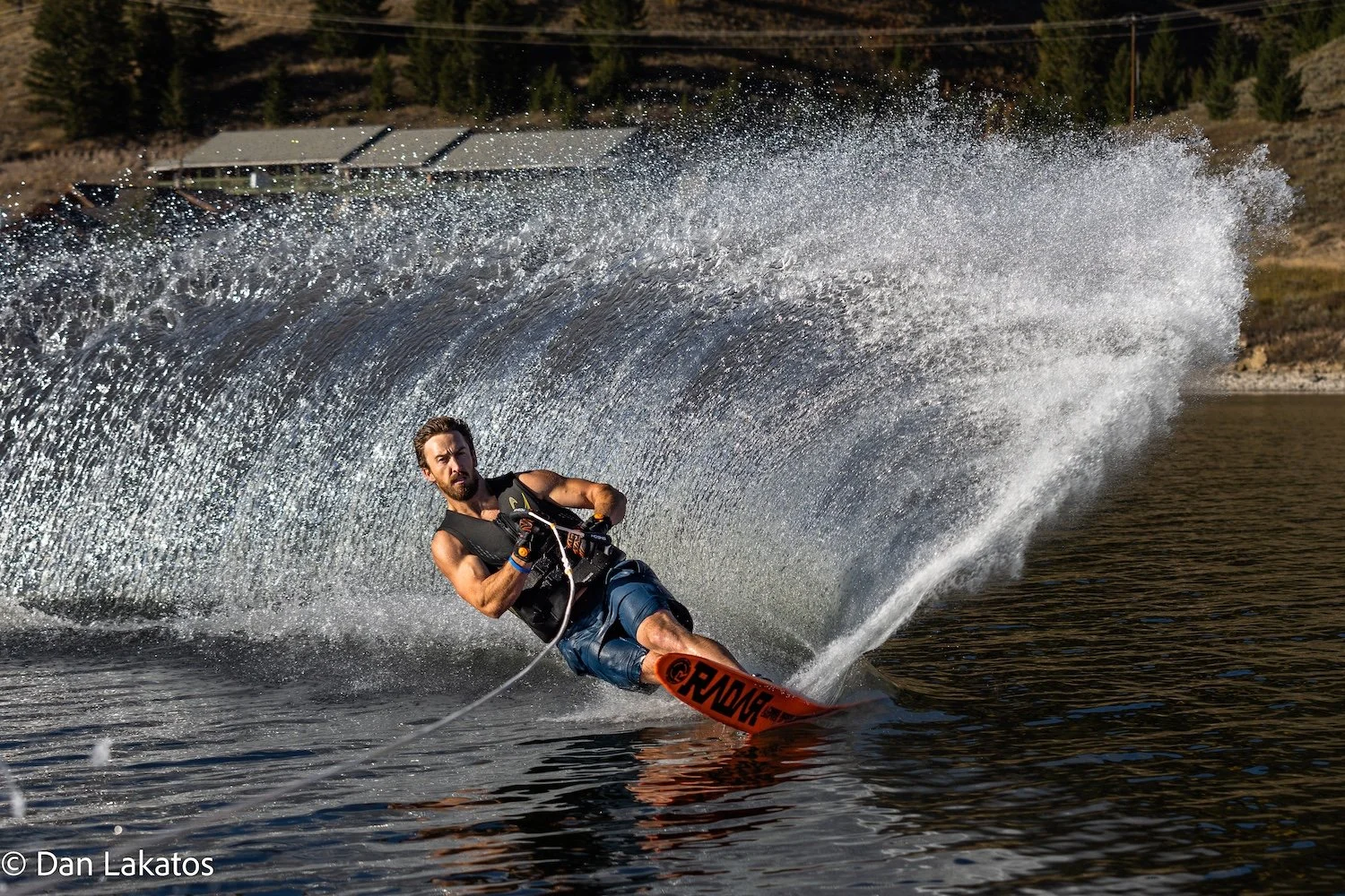 A man waterskiing on a lake, making a sharp turn and creating a large wake with water spray behind him, in a natural outdoor setting. Hebgen Lake, Montana, USA