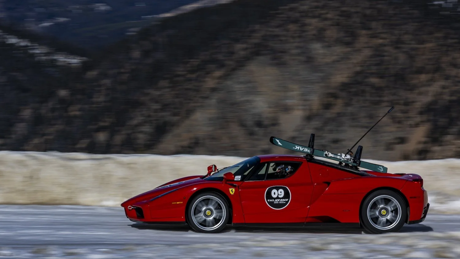 A red Ferrari race car with the number 09 on the side, equipped with skis on the roof, driving on an icy track with snow and mountains in the background. F.A.T. ICE RACE 2026, Big Sky, Montana USA. Dan Lakatos Photo
