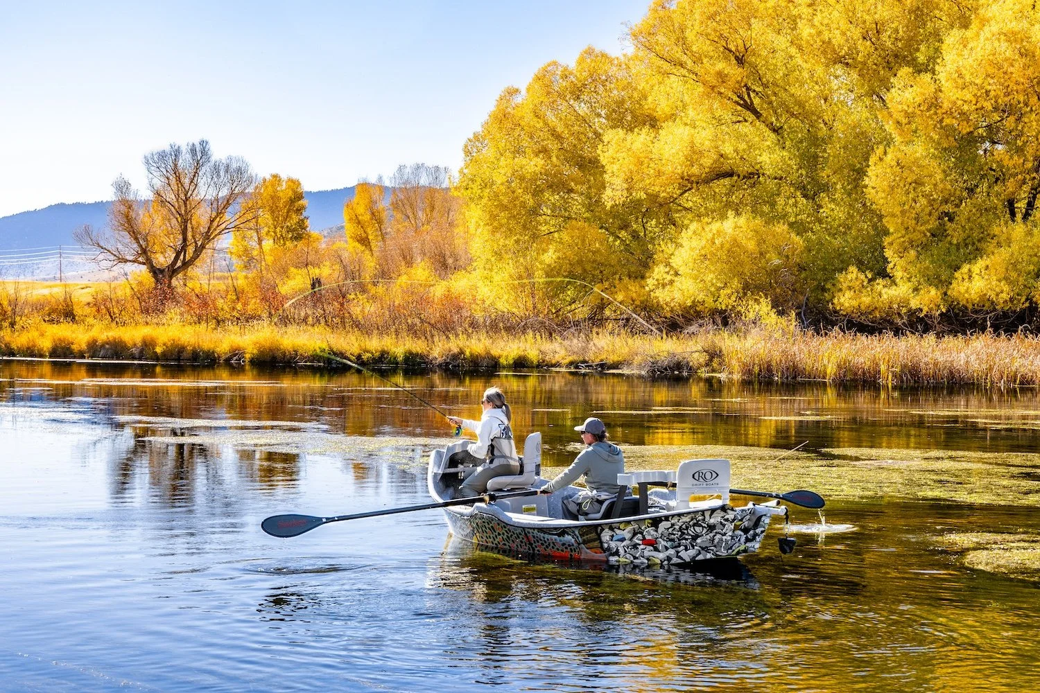 Two people fishing on a boat in a river with autumn trees and clear sky in the background. Livingston, Montana, USA
