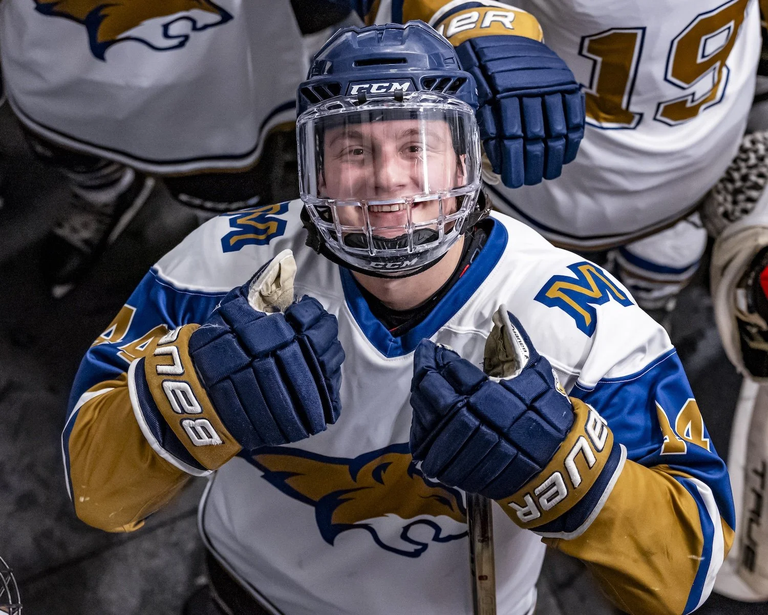 A hockey player in a white, blue, and gold jersey with the number 19 and a lion logo, wearing a helmet and gloves, smiling and pointing at himself in a hockey arena.