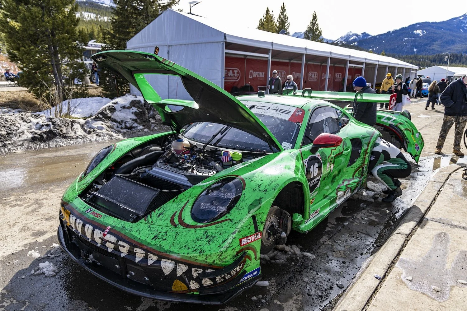 Green race car with open hood, parked on a paddock area with snow on the ground. There are people working on the car and others standing nearby, with mountains and a tent in the background. F.A.T. ICE RACE 2026, Big Sky, Montana USA. Dan Lakatos Phot